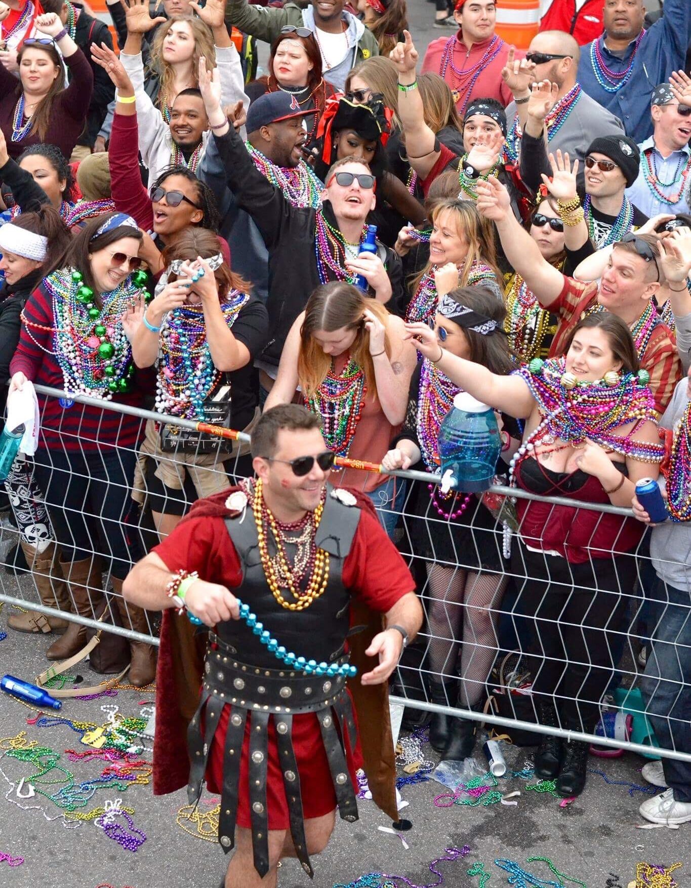 Crowd at Gasparilla Pirate Fest wearing beads and cheering, with a Roman soldier handing out beads in the foreground.