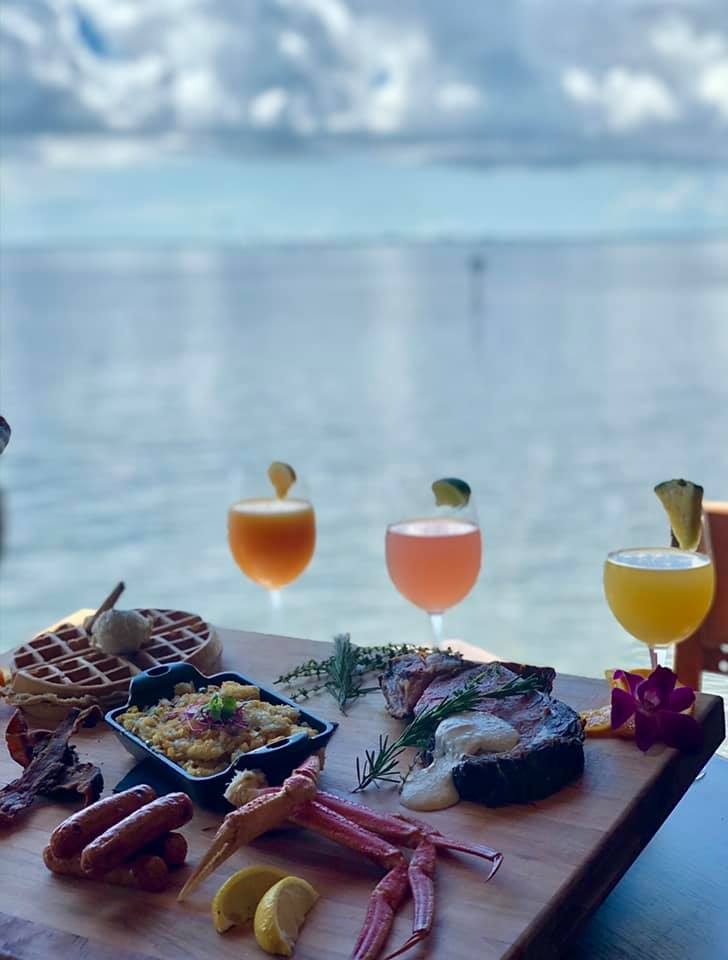 Image of three cocktails and a variety of brunch entrees by the water during brunch at The Rusty Pelican