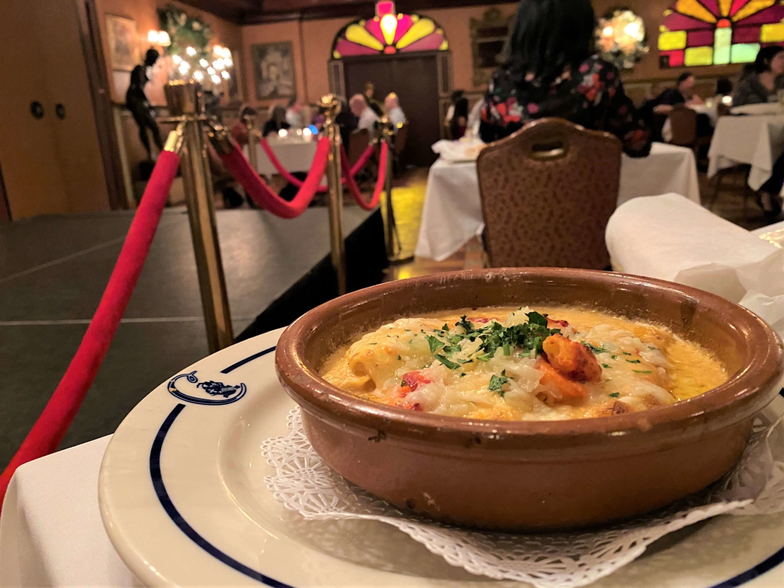 Cannelloni de Langosta Columbia Ybor City is in the foreground served in a circular clay dish. The dining room in the background is dimly list with red and yellow stained glass windows at the very back wall