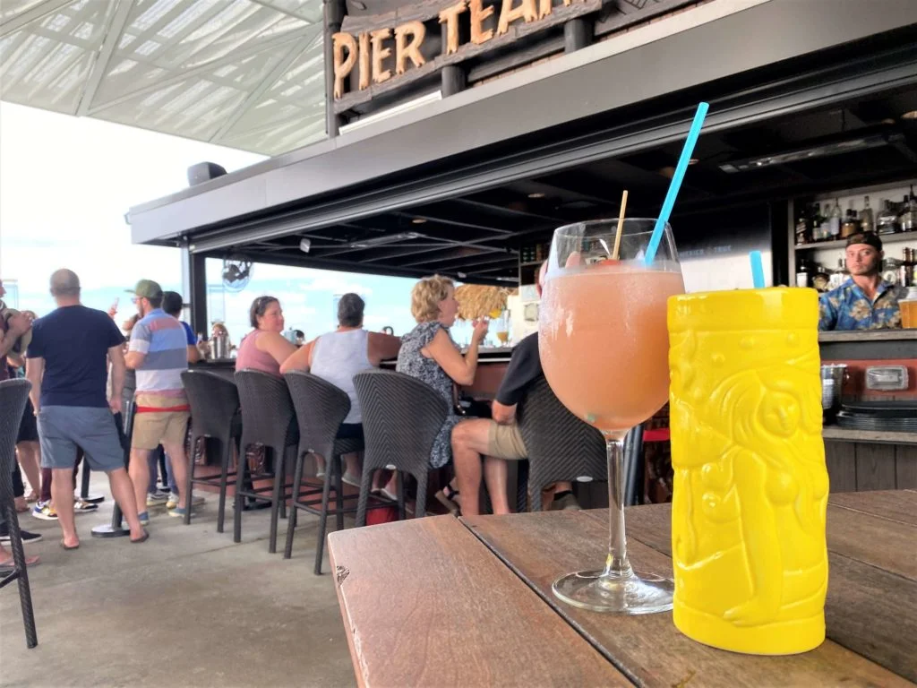 Tropical Drinks at Pier Teaki St. Pete include a tall bright yellow tiki mug with a mermaid scene on it, and light pink colored frozen rose wine drink, sitting on a dark wood table in the foreground and the outdoor bar in the background