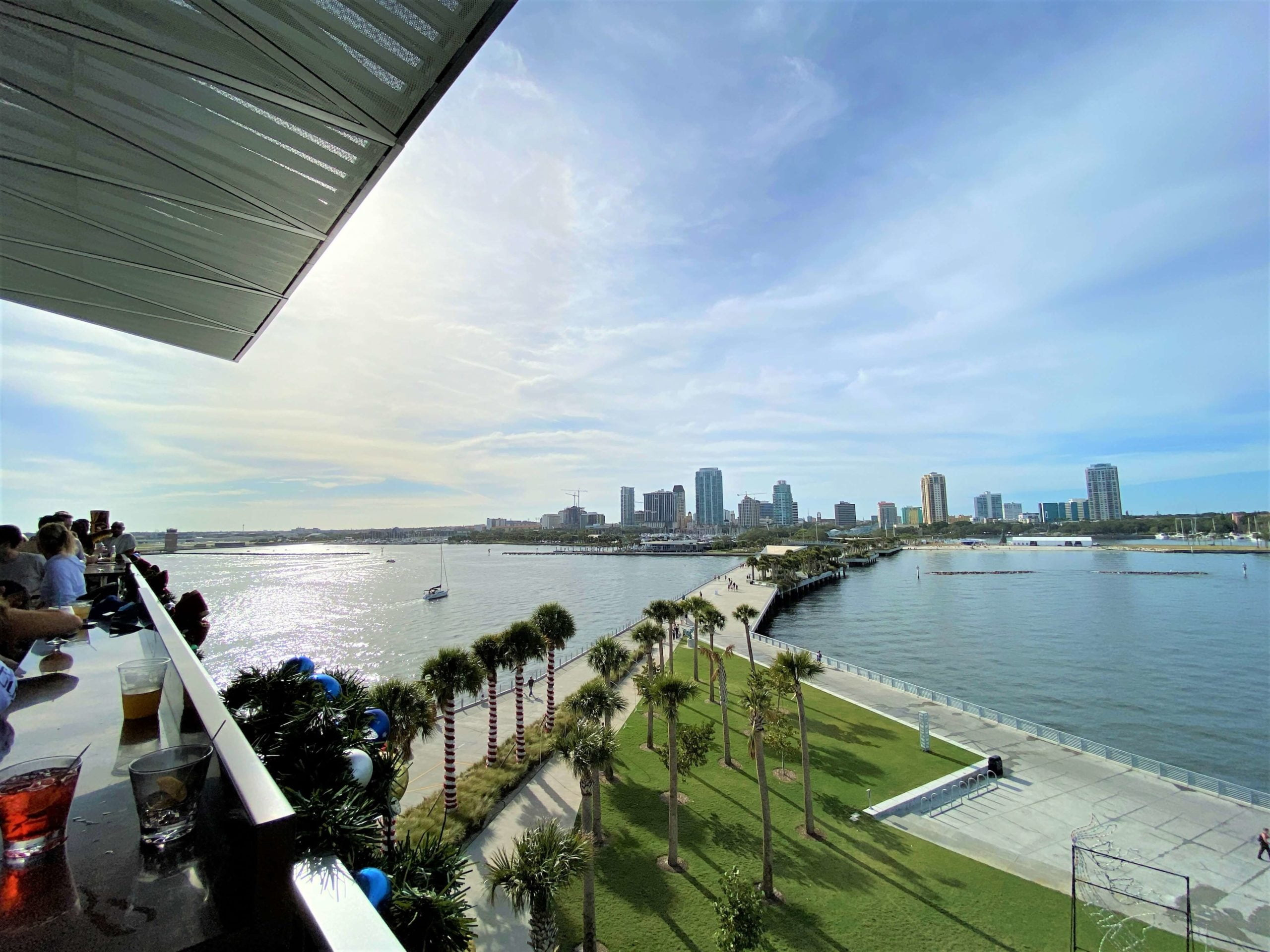 View of St. Pete Pier from Pier Teaki includes a green lawn and palm trees in the foreground, the long St. Pete pier in the middle, and the city scape of St. Petersburg Florida in the background.