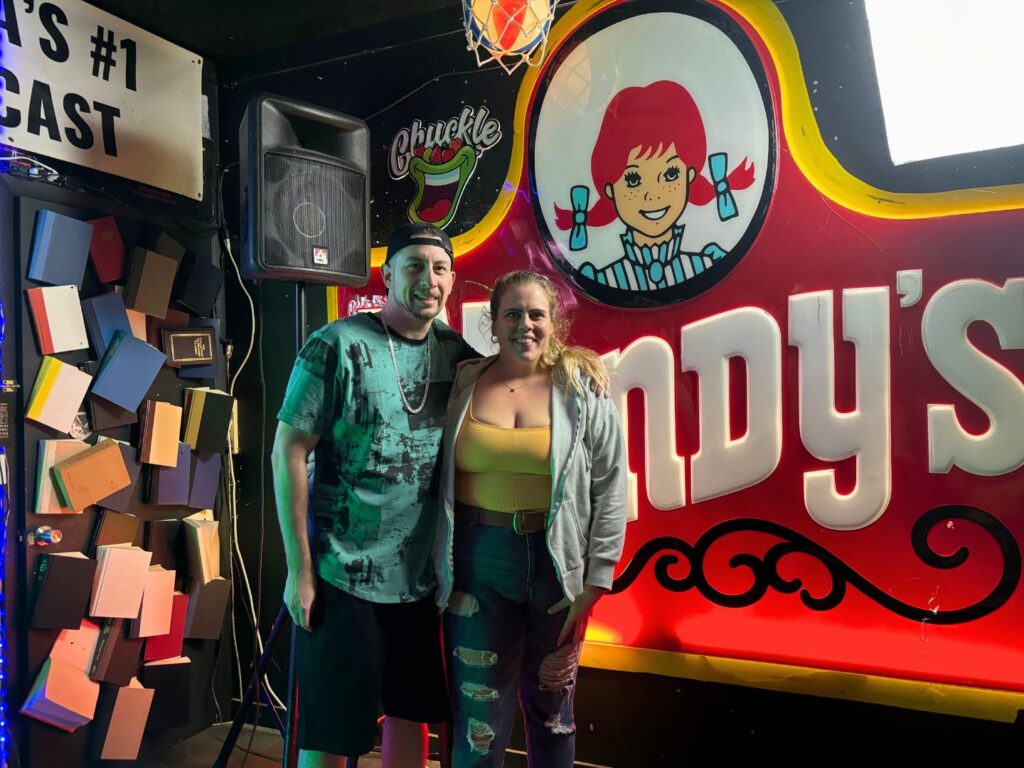 a couple poses in front of props and signs at The Gimmick comedy club in Ybor City