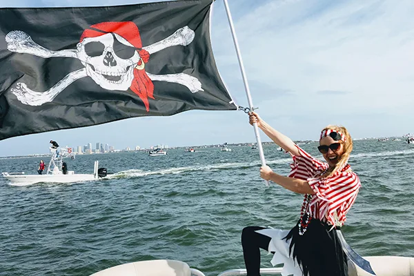 Smiling woman dressed as a pirate holding a Jolly Roger flag on a boat during the Gasparilla Invasion in Tampa Bay.