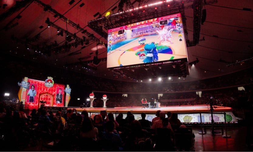 A virtual Pokémon battle happening on a big stage with a screen showing the players in battle