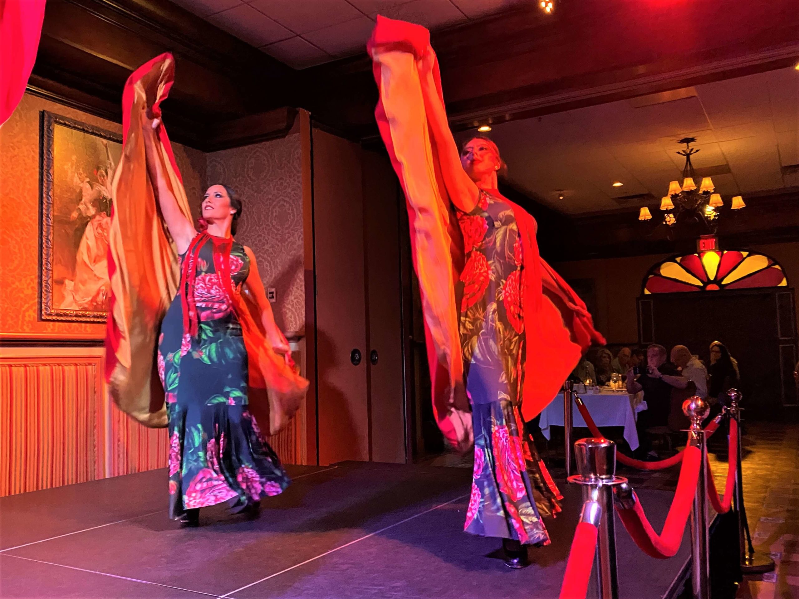 Two costumed dancers are on stage at Flamenco Dance Show at Columbia Restaurant Ybor City wearing long dark dresses and have their arms raised as they perform with red velvet capes